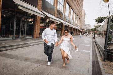 Wedding photography, portrait of happy smiling newlyweds, stylish groom and beautiful bride, walking, running barefoot with shoes in hands and holding hands, outdoors on the street in the city.