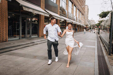 Wedding photography, portrait of happy smiling newlyweds, young groom and beautiful bride, walking, running barefoot with shoes in hands and holding hands, outdoors on the street in the city.