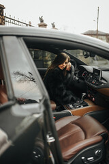 A portrait of a beautiful, stylish young woman, a female model in a fur coat and lingerie, sitting and posing inside, behind the wheel of an expensive, luxury premium car. Advertising shoot.