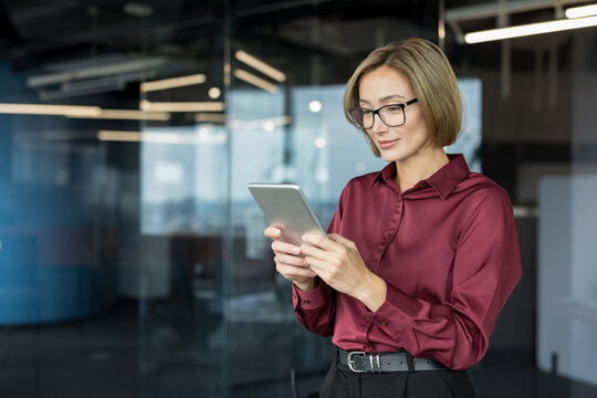 Young businesswoman with glasses researching on a digital tablet, standing in a contemporary creative office with glass walls, representing technology integration in professional work - Powered by Adobe