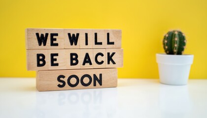 Wooden blocks spell We Will Be Back Soon. Small potted cactus sits nearby. White surface with bright yellow background. Business reopening message concept.