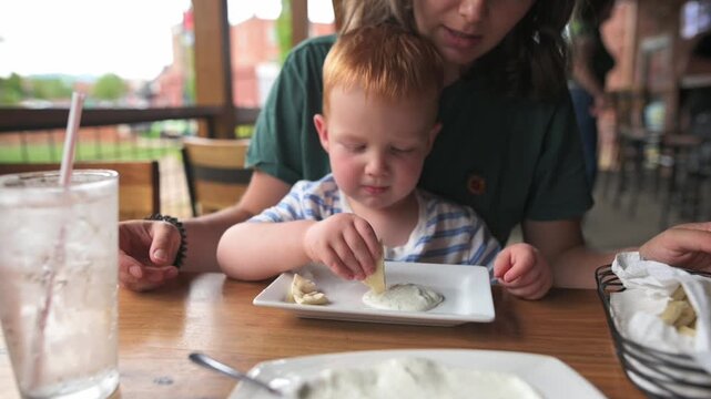 caucasian toddler tasting new food at outdoor restaurant, child pokes and samples small bites while parent looks on, facial reaction and tiny fingers on plate create authentic tasting moment