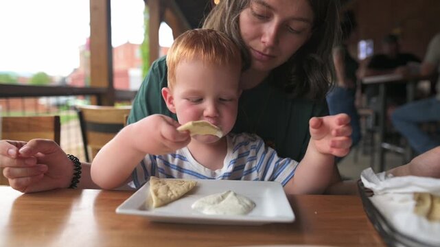 toddler hesitates prior to tasting crunchy chips, mother gently guides her child as he attempts snack tasting, mother softly supports her young child during process of trying his first snack