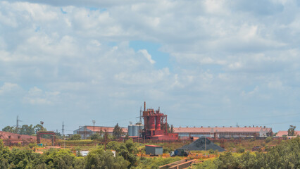 Obraz premium Industrial landscape featuring a red furnace and factory buildings, surrounded by green trees under a bright, cloudy sky