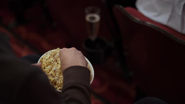 CloseUp Popcorn In Hand Dim Cinema Seat With Red Upholstery And Glass In Holder, Relaxed Viewer Reaches Into Bowl, Fingers Picking Kernels,