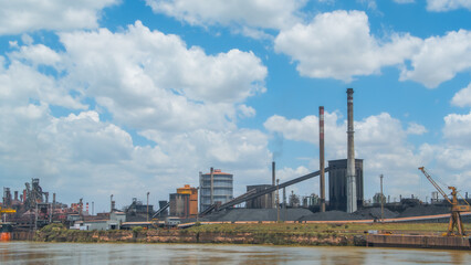 Obraz premium Industrial steel mill or factory complex with tall smokestacks, large conveyor belts, and piles of coal on a riverside under a vast cloudy blue sky