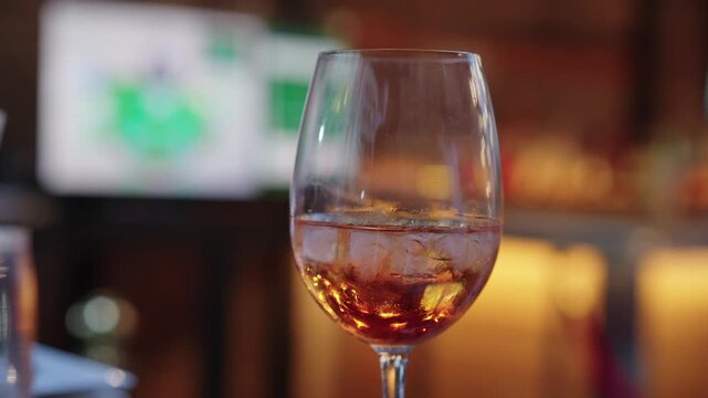 Cocktail Glass On Bar With Television, Amber Liquid Swirling, Condensation On Stemware, Warm Bokeh Lights, Sports Broadcast Blurred, Relaxed