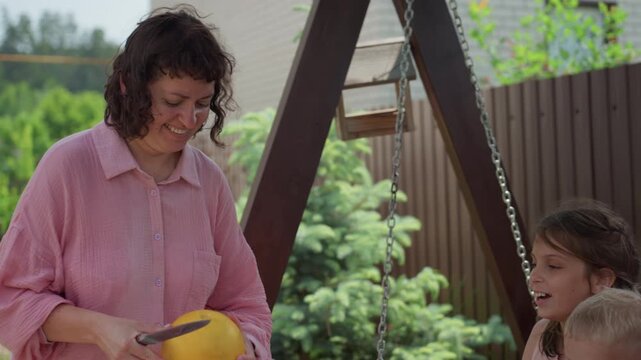 Caucasian Woman Entertains Children With Melon And Rhythm In Garden, Kids Clap And Cheer At Picnic Table Near Swing, Wooden Spoon Rhythm Game Creates Joyful Family Bonding In Sunny Backyard