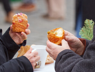An older couple enjoys sweet filled puff pastry and hot drinks at a table at Naplavka farmers market, wrapped in winter coats, capturing the warm Advent atmosphere of the winter season in Prague.