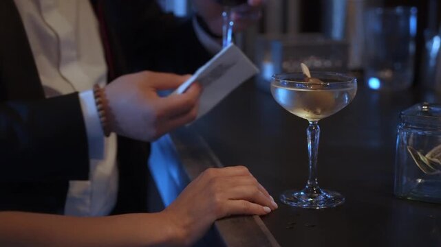 Hands Exchanging Coupe Glass Over Bar Counter, Suit Cuff And Female Wrist Visible, Tip Jar With Cash On Side, Soft Blue Lighting, Quiet