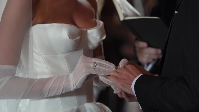Bride Sliding Ring Onto Groom Finger During Vows, Gloved Hands And Satin Dress Visible While Officiant Holds Book Nearby Creating Solemn,