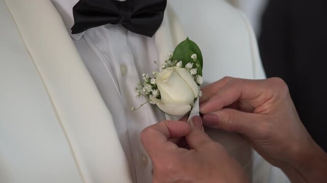 Groom Tux Boutonniere Being Carefully Pinned To Lapel, White Rose With Delicate Filler Flowers Placed By Attentive Hands, CloseUp Of Bow