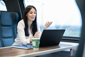 A cheerful woman engages in a video call using her laptop in a train, beautifully blending work and travel moments, showcasing the modern lifestyle of digital nomads.