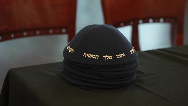Navy Kippah Stacked On Tablecloth, Wooden Chairs Blurred Background, Gold Hebrew Embroidery Catching Soft Light, Stacked Kippot Suggesting