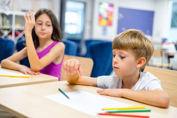 Fototapeta premium Two kids in a modern classroom drawing and raising hands during a lesson. Concept of active learning, creativity, early education, and smart school