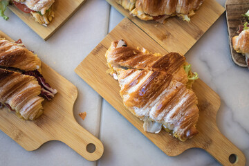 Top view of several freshly made croissant sandwiches served on small wooden cutting boards on a light marble table