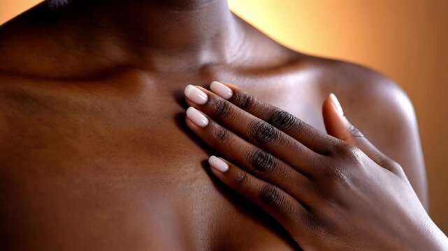 Close up of a dark-skinned woman's hand with light pink manicured nails gently touching to her collarbone and chest area