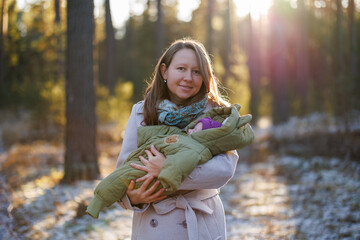 A girl with a child in a sunny winter forest. A mother holds her daughter in a park. The woman smiles at her newborn. The baby is wearing a winter jacket.