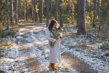 A girl with a child in a sunny winter forest. A mother holds her daughter in a park. The woman smiles at her newborn. The baby is wearing a winter jacket.