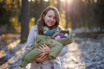A girl with a child in a sunny winter forest. A mother holds her daughter in a park. The woman smiles at her newborn. The baby is wearing a winter jacket.