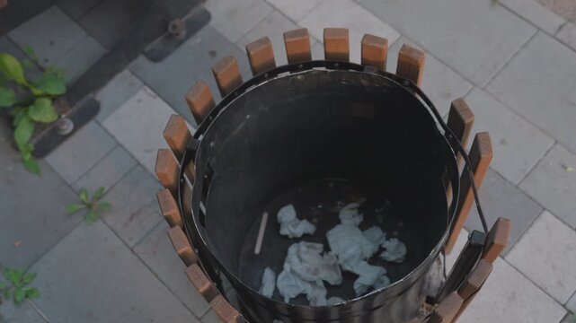 Person Drops Tissue Into Bin, Overhead Shot Of Hand Releasing Crumpled Tissues Into Wooden Waste Container On Tiled Pavement, Nearby Plant And Green Edging Visible, Deliberate Ritualized Motion