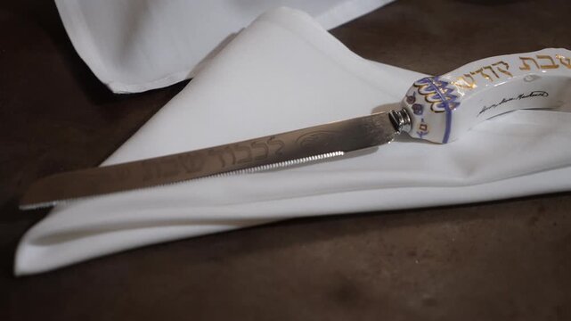 White Napkin With Ceremonial Challah Knife, Ceramic Handle Embossed Hebrew Inscription Resting On Folded Cloth Over Dark Table, Soft Warm