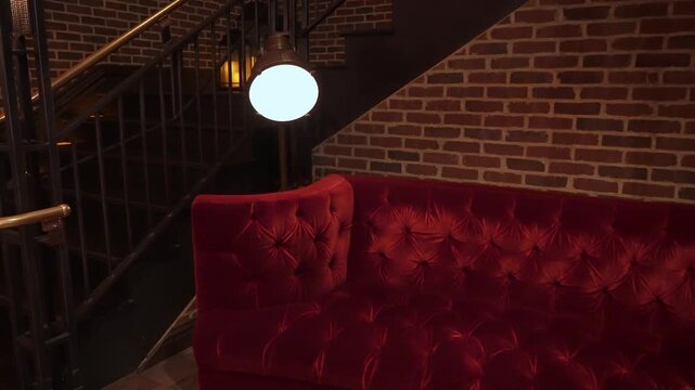 Red Velvet Sofa Under Lamp, Tufted Booth Framed By Exposed Brick And Iron Staircase, Warm Ambient Lighting, Plush Seating For VIP Guests,