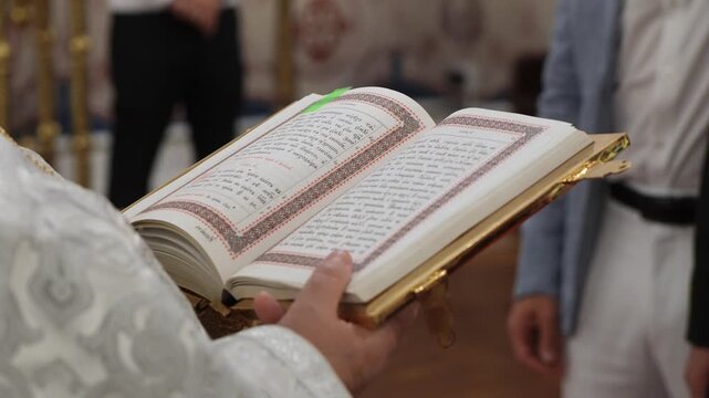 Open Gilded Book Held By Officiant In Solemn Liturgical Ceremony, Embroidered White Robe, GoldEdged Pages, Hands Turning Folio, Blurred