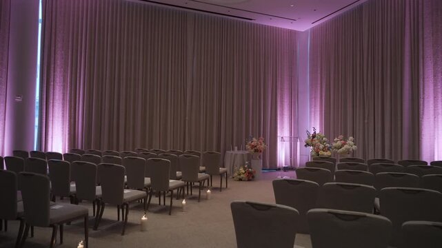 Aisle Lined Chairs With Purple Uplighting And Candles Leading To Floral Altar Empty Boutique Ballroom Framed By Heavy Drapery, Soft