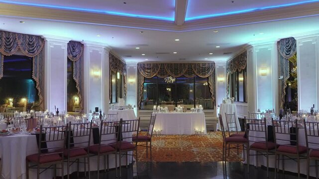 Empty Hotel Ballroom With Blue Uplighting, Round Tables Dressed In White Linen, Chiavari Chairs, Elevated Head Table With Backdrop, Polished