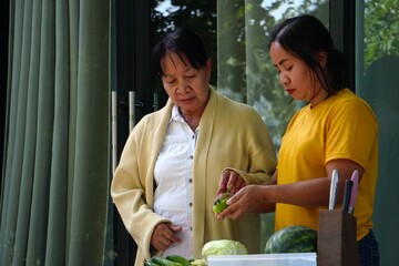 The older woman watches her daughter who is peeling a cucumber in preparation for cooking.