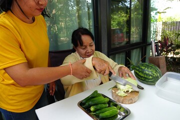 A woman in a yellow shirt holds her mother's arm to help stabilize her while cutting cabbage.