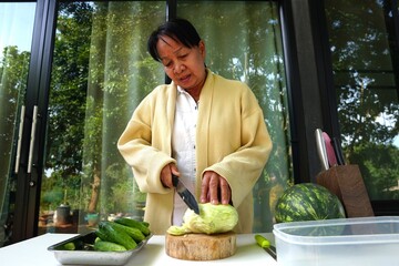 The older woman is concentrating on using a knife to slice cabbage on a wooden chopping board.