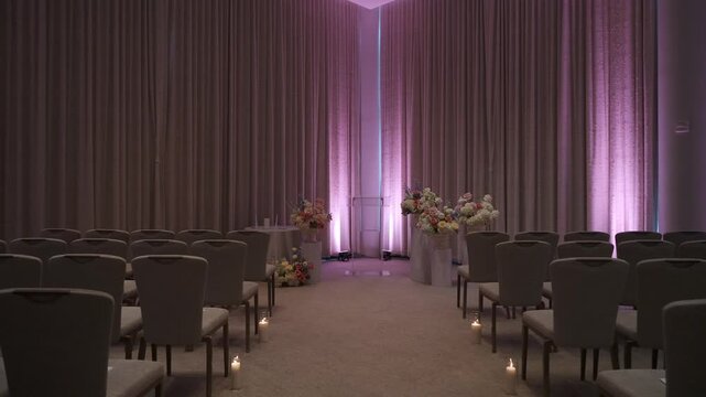 Aisle Lined Chairs With Purple Uplighting And Candles Leading To Floral Altar Empty Boutique Ballroom Framed By Heavy Drapery, Soft