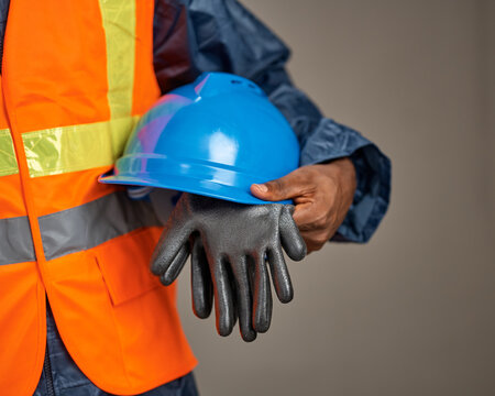 A close-up shows a worker wearing an orange reflective safety vest holding a blue hard hat and a pair of protective gloves against a neutral studio background.