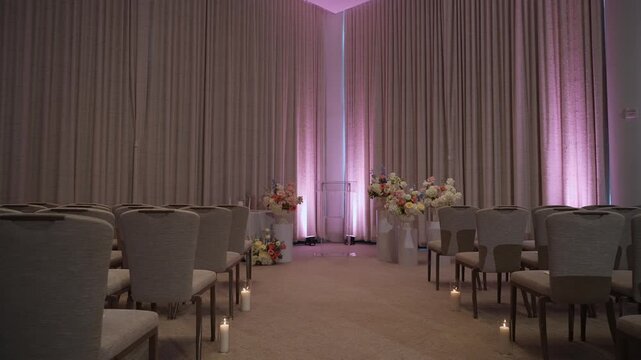 Aisle Lined Chairs With Purple Uplighting And Candles Leading To Floral Altar Empty Boutique Ballroom Framed By Heavy Drapery, Soft