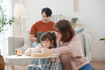 A cheerful Asian mother helps her young daughter do homework and color a book at home, while the father works on a laptop in the background. A cozy, bright, and modern family lifestyle.