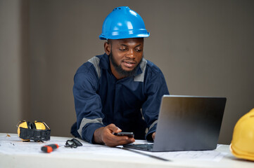 A young Black man wearing blue industrial coveralls and a blue safety helmet leans over a table using a laptop while holding a smartphone, with tools and safety gear beside architectural plans.