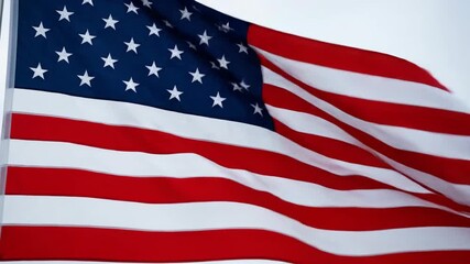 A bright, patriotic close-up shot captures the iconic red, white, and blue american flag dramatically waving and fluttering against a clear sky background.