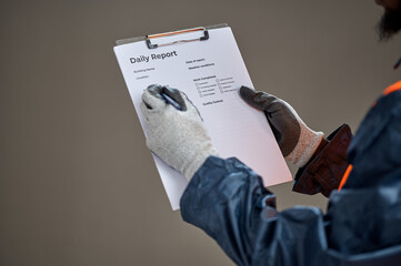 Fototapeta premium Close-up of an African adult construction worker wearing protective gloves and safety vest while writing on a daily report clipboard during a workplace inspection against a plain background.
