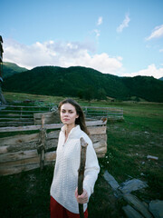 Fototapeta premium Woman in a white knitted sweater stands beside a wooden fence in a wide countryside field, carrying a wooden stick, with green hills and blue sky behind