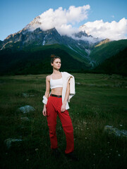 Fototapeta premium Mountain landscape with a stylish woman standing in a green meadow, wearing a white crop top and red pants, jacket draped over shoulder, crisp sky and distant peaks.