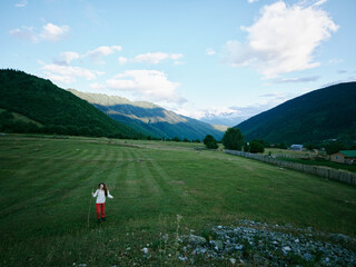 Fototapeta premium Wide meadow between hills with a girl walking across green grass, distant mountains under a bright sky and clouds, capturing freedom, nature, and outdoor exploration