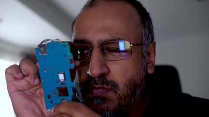 South Asian IT engineer is inspecting a motherboard with a loupe. He is repairing computer hardware and checking the circuit board for damage performing electronics maintenance.