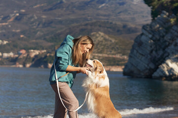 A Border Collie stands beside a woman in a teal jacket on the rocky beach. They enjoy a quiet moment near calm blue waters.