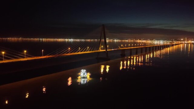 Lit catamaran navigates dark waters of Paran&aacute; River beneath the illuminated cables of the San Roque Gonz&aacute;lez de Santa Cruz Bridge connecting Argentina and Paraguay at night, drone tracking and orbit