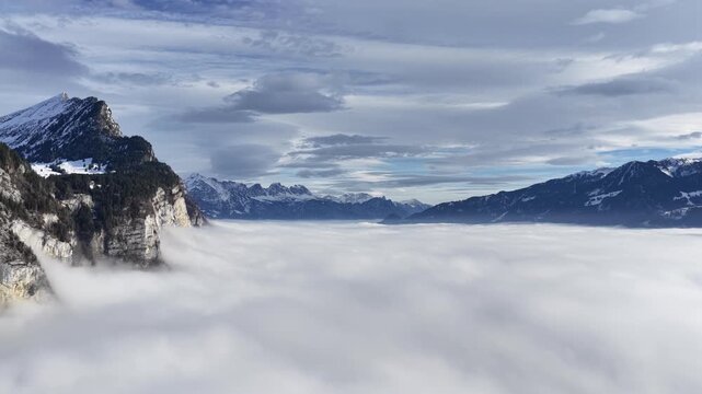 High-altitude view over a dense fog inversion in Amden, Switzerland. The Swiss Alps and Churfirsten peaks emerge from the vast, rolling clouds. An inspiring aerial scene of natural escape