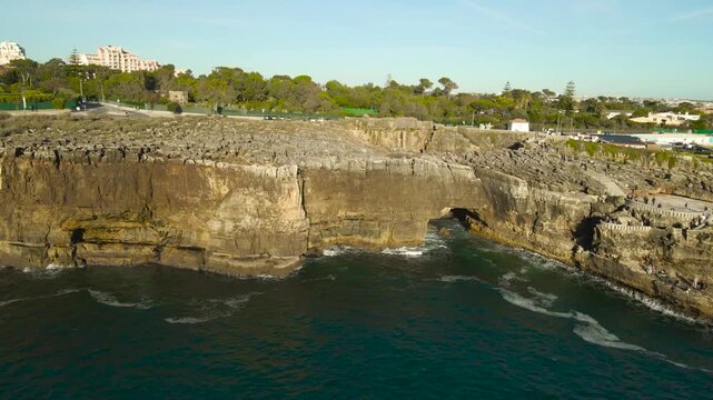 Aerial drone footage orbiting and flying around a famous tourist attraction on Portugal cliff bank coast shoreline called Boca do Inferno or Devils mouth where tourists are watching it on platforms.
