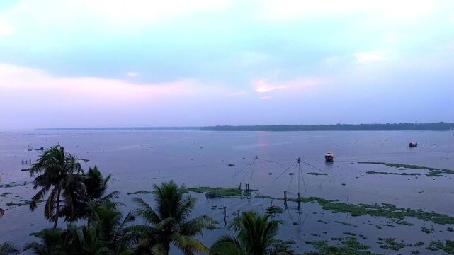 Aerial sunset view of Kerala backwaters and Vembanad Lake at dusk, featuring Chinese fishing nets, palm trees, and a distant houseboat creating a serene, purple landscape.