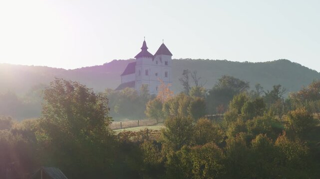 Aerial shot a Herina, Bistrita Nasaud, rRomania white citadel church nestled on a hill surrounded by trees and mist.